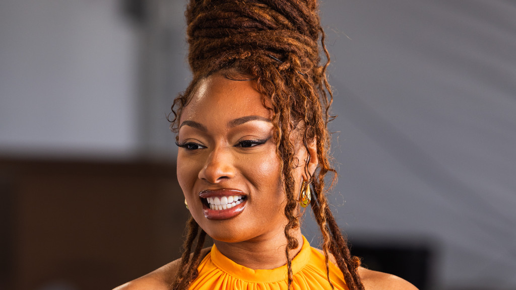 A radiant woman with reddish-brown dreadlocks styled in an updo smiles widely, showing white teeth, and wears a bright orange top and a gold earring.