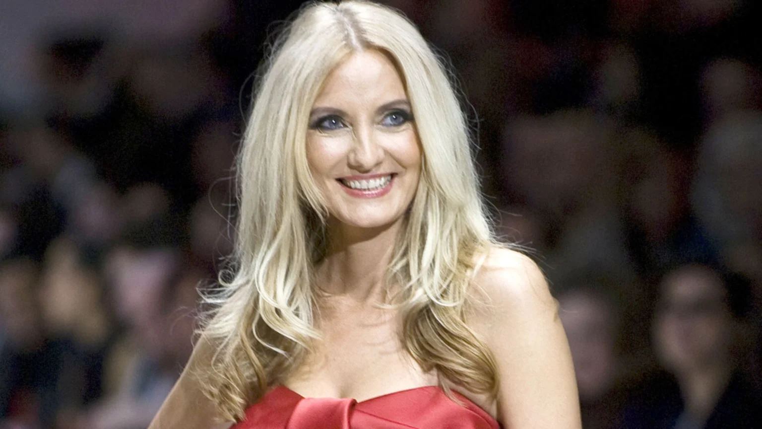 A smiling blonde woman with long wavy hair, blue eyes, and dark eyeliner is wearing a strapless red top against a blurry dark background with indistinct figures.