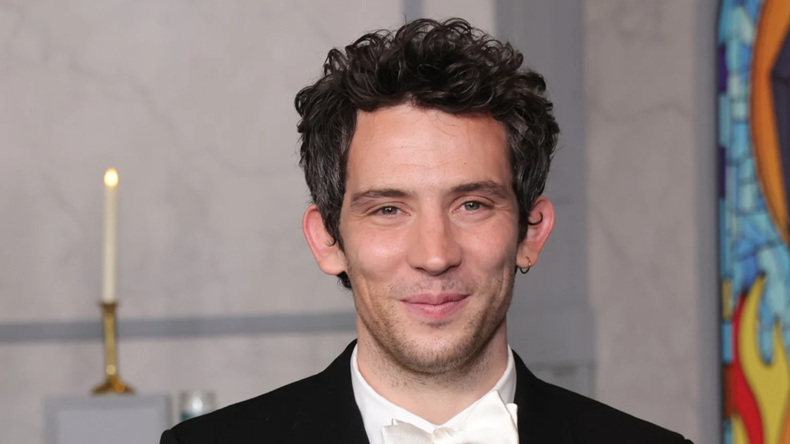 A smiling man with curly dark hair, light eyes, a slight beard, and a small hoop earring wears a black suit and white bow tie, with a lit candle and colorful art in the soft-focus background.