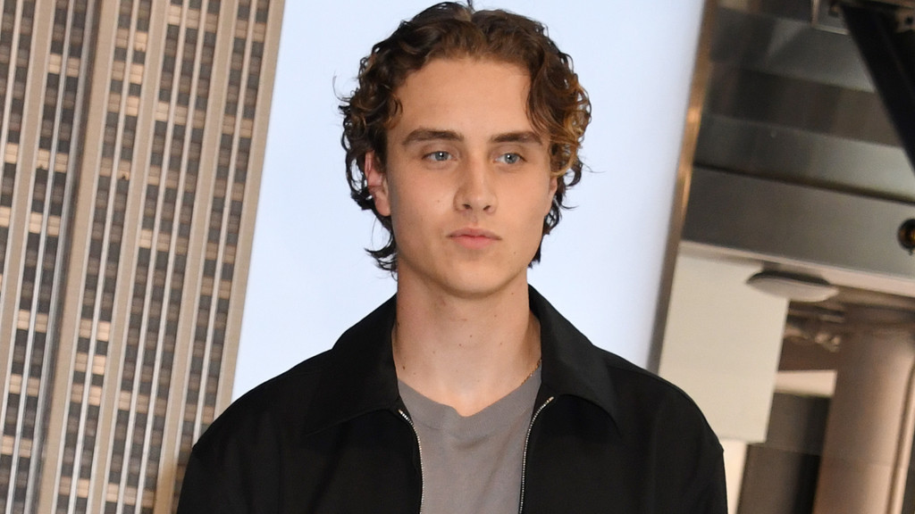 A young man with curly brown hair and blue eyes, wearing a black collared jacket over a grey t-shirt, looks directly at the camera with a neutral expression, with a skyscraper visible in the background on the left.