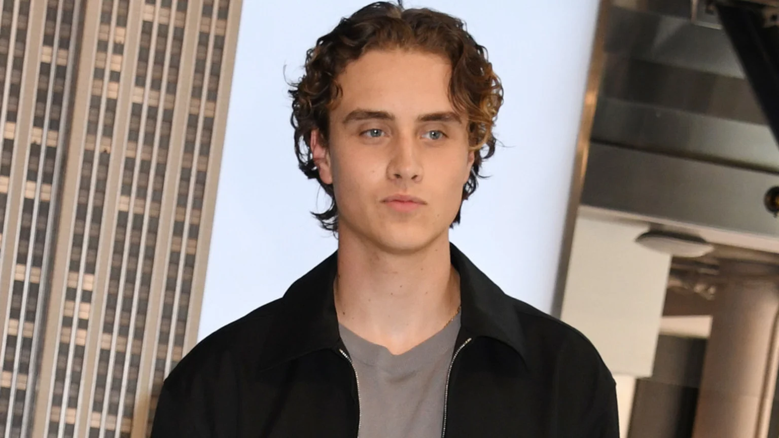 A young man with curly brown hair and blue eyes, wearing a black collared jacket over a grey t-shirt, looks directly at the camera with a neutral expression, with a skyscraper visible in the background on the left.