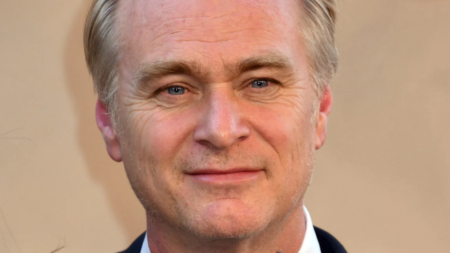 Close-up of a smiling middle-aged man with fair skin, blue eyes, and blondish-grey hair, wearing a dark suit and white shirt.