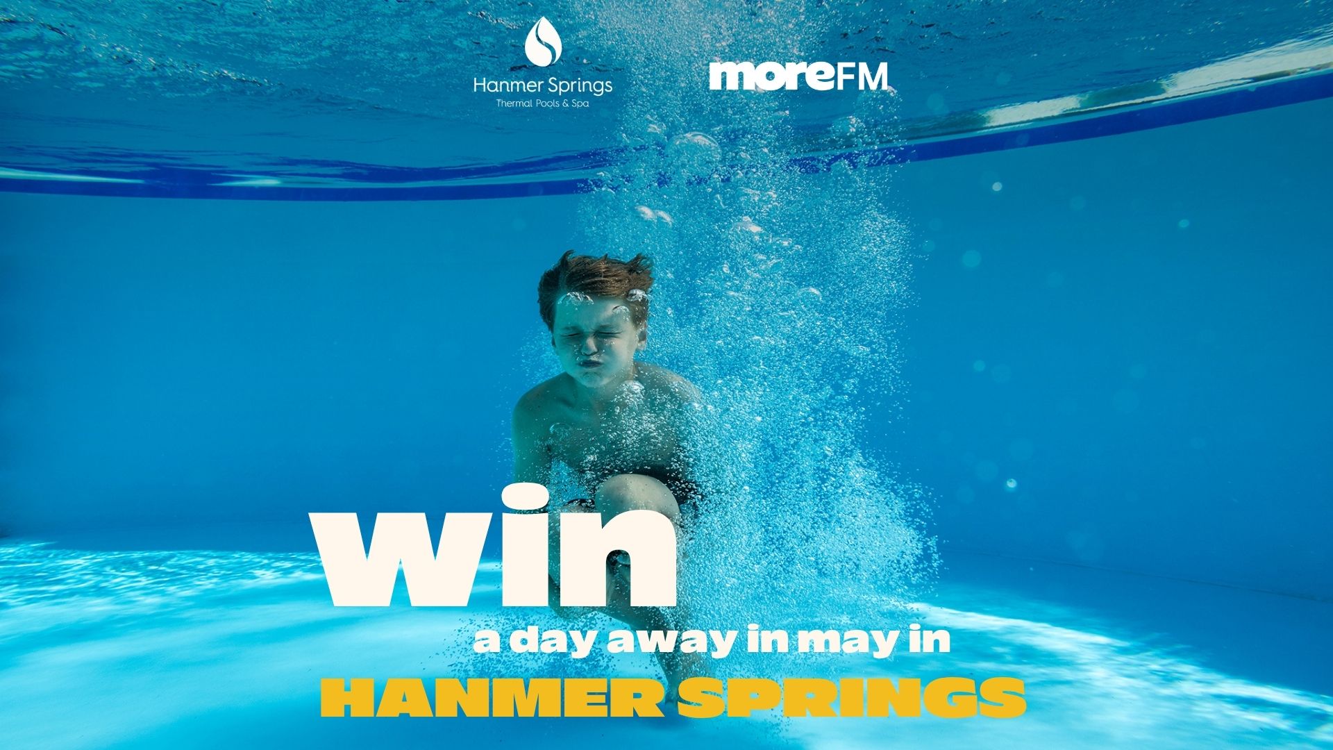 Underwater shot of a young boy in a blue pool with bubbles, overlaid with text "win a day away in May in HANMER SPRINGS" and logos for Hanmer Springs and moreFM.