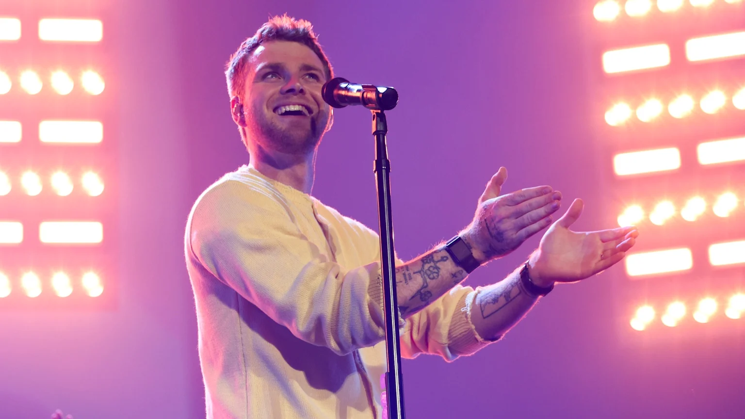 A smiling man on stage, wearing a light sweater and with visible arm tattoos, looks up while gesturing with open hands in front of a microphone, with bright stage lights in the background.