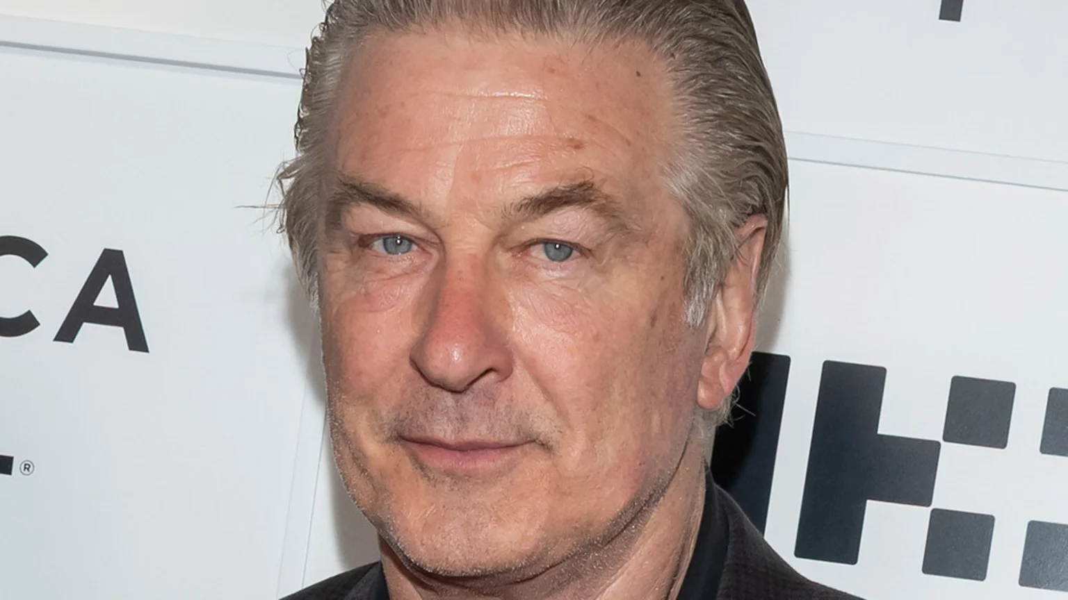 A close-up headshot of actor Alec Baldwin, with grey hair and blue eyes, looking at the viewer with a neutral expression against a white background with black text.