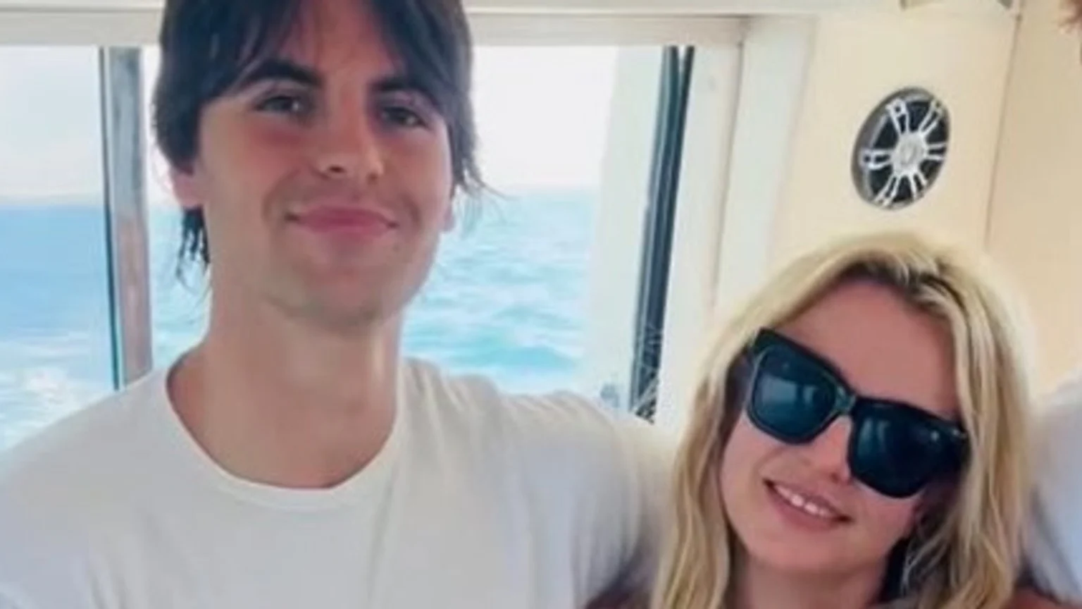 A young man with dark hair and a blonde woman in black sunglasses smile at the camera, sitting together on what appears to be a boat with the ocean visible through windows behind them.