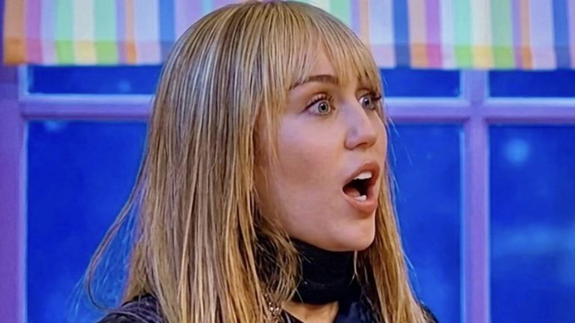 Miley Cyrus, with long blonde hair and bangs, wears a cream embroidered jacket and sequined top, pressing a finger with rings to her lips in a "shhh" gesture, against a blurry pink background with soft lights.