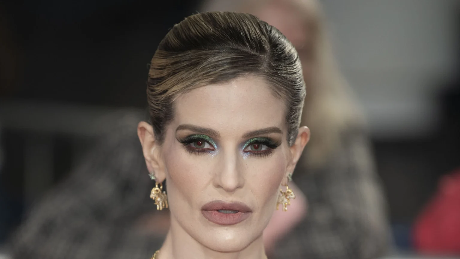 A woman with light brown hair pulled back, wearing dramatic green eyeshadow, glittery blue under-eye makeup, dark eyelashes, nude lipstick, gold ram-shaped earrings, and reddish eyes.