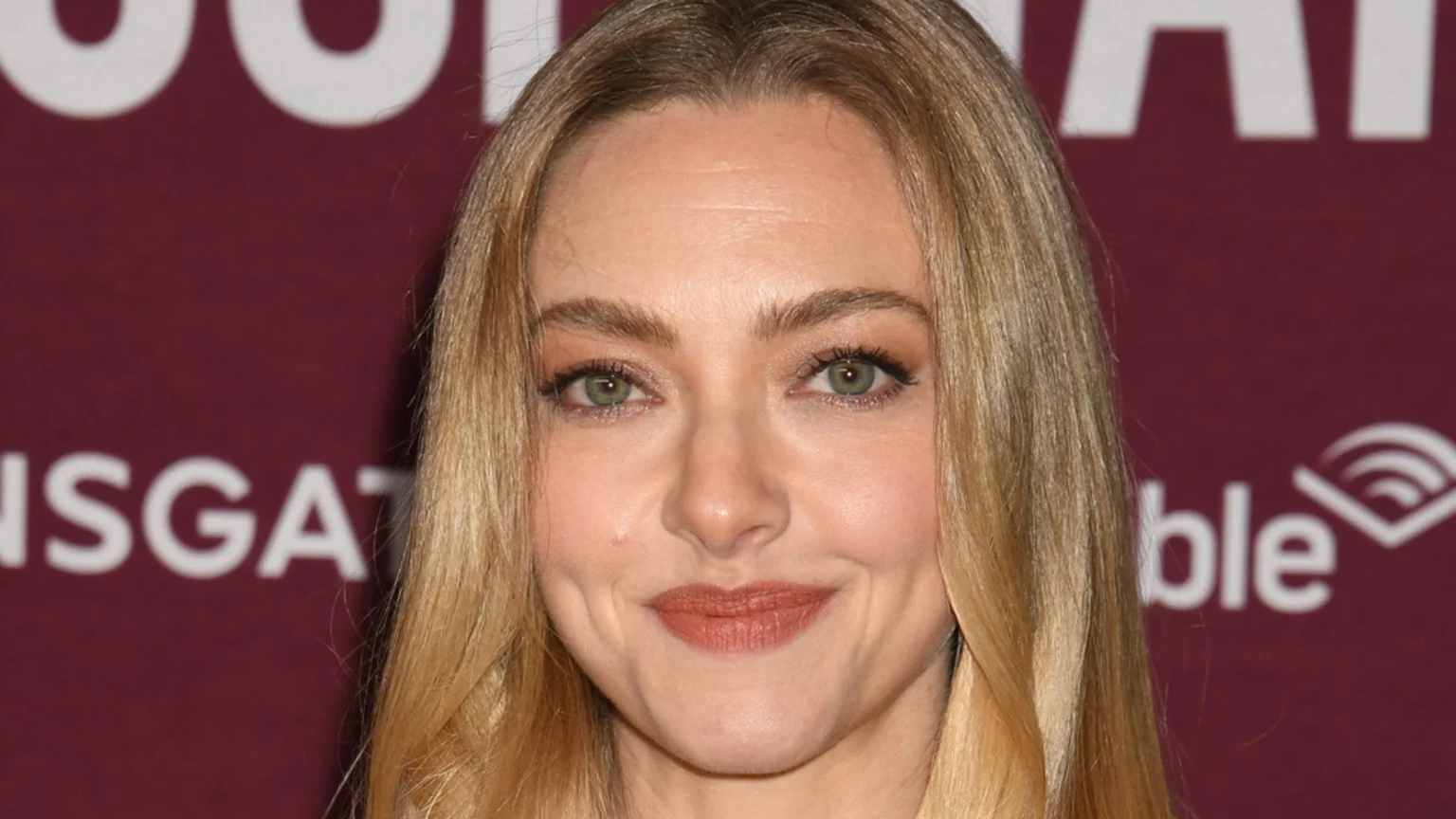 Close-up of actress Amanda Seyfried smiling slightly, with blonde hair and green eyes, against a blurred dark red background featuring parts of the Lionsgate and Audible logos.