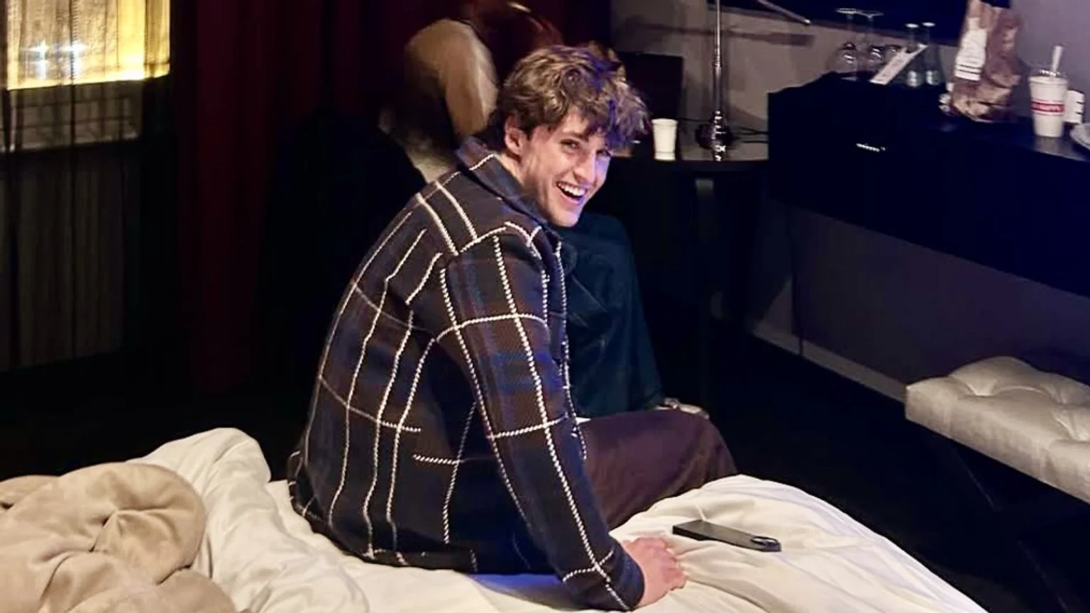 A smiling young man with messy brown hair and a plaid jacket looks over his shoulder while sitting on a bed in a hotel-like room with a lamp and dark furniture.