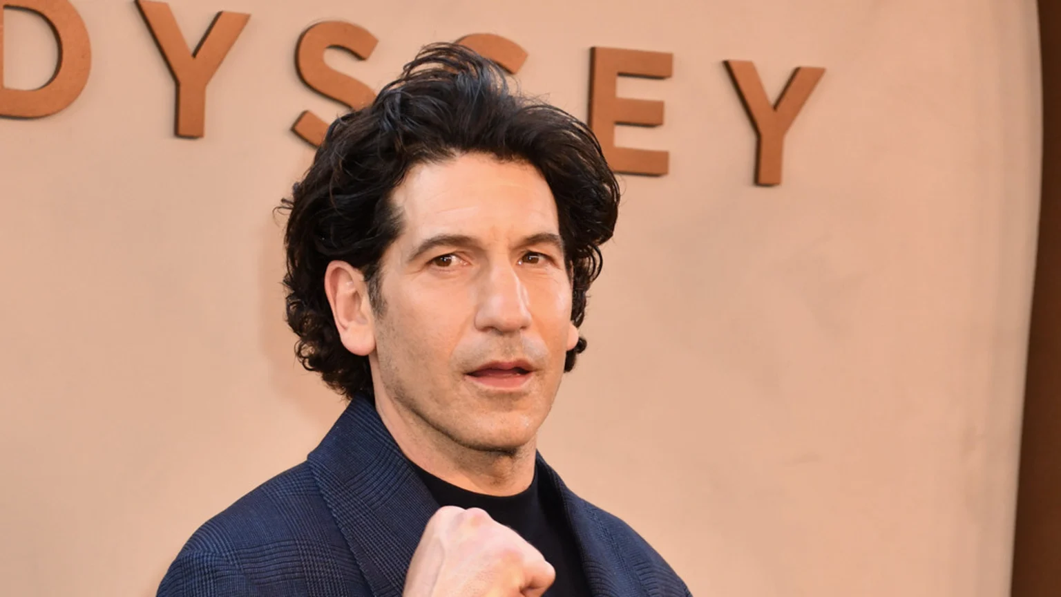 Jon Bernthal, with dark curly hair and a plaid jacket, holds a clenched fist in front of a beige wall with brown "ODYSSEY" letters.