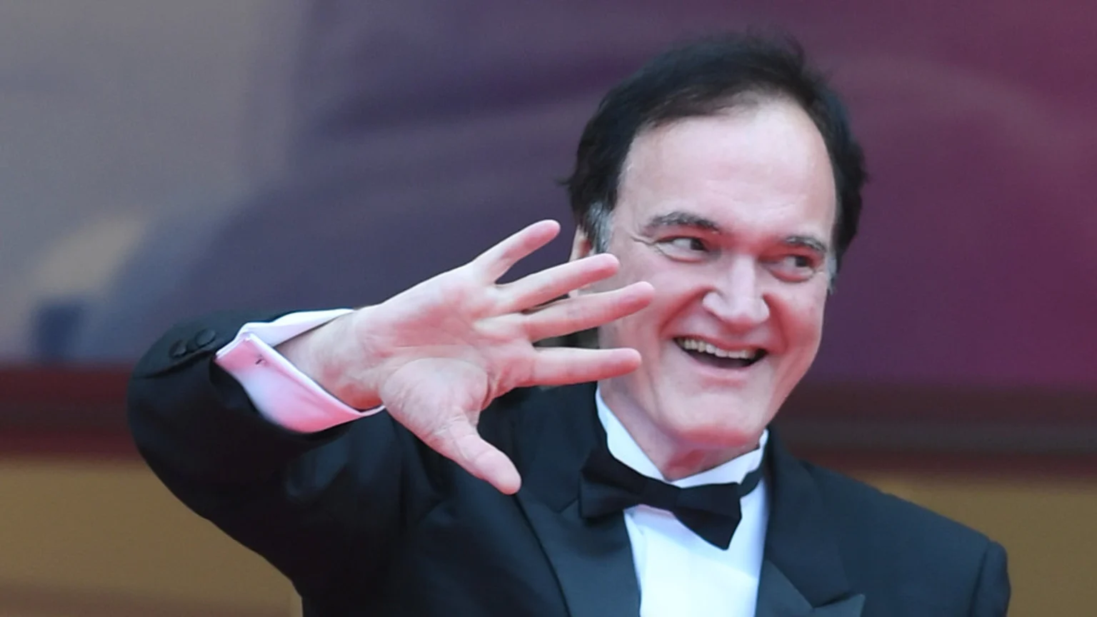 Waving and smiling director Quentin Tarantino, wearing a black tuxedo and bow tie, looks to his right with his left hand raised, palm forward.