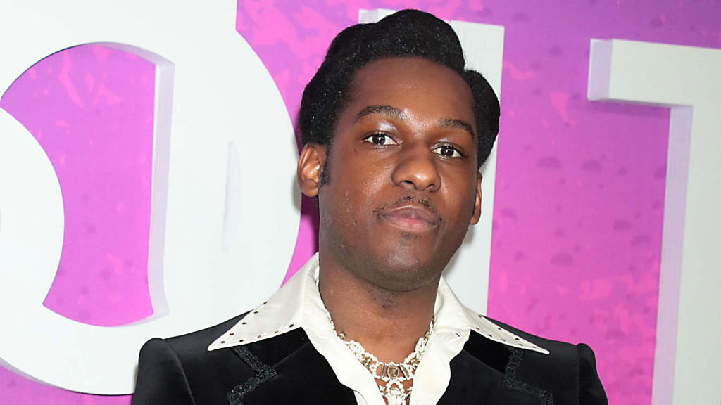 A Black man with a pompadour hairstyle and mustache wears a black suit, white collared shirt, and necklaces, looking at the camera against a pink and white background.