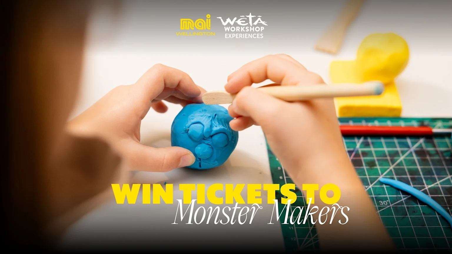A child's hands use a sculpting tool to shape a blue clay monster face, with a green cutting mat, yellow clay, "mai WELLINGTON" and "WĒTĀ WORKSHOP EXPERIENCES" logos, and "WIN TICKETS TO Monster Makers" text.
