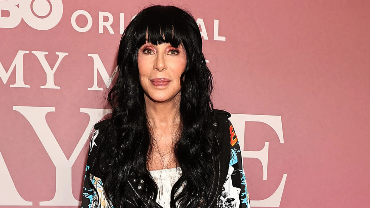 Cher, with long black hair, bangs, and pink eyeshadow, wears a black patterned jacket and white top, looking forward against a pink wall with white "HBO" text.