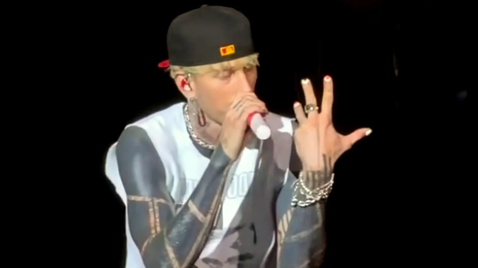 Rapper Machine Gun Kelly on stage, wearing a black cap and white shirt, sings into a mic, raising his tattooed left arm with rings, bracelets, and multi-colored nail polish.