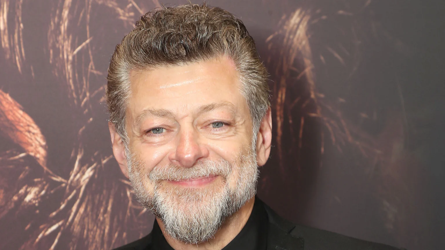 A smiling Andy Serkis with grey hair and beard, wearing a black shirt and jacket, against a dark, abstract background with brown tones.