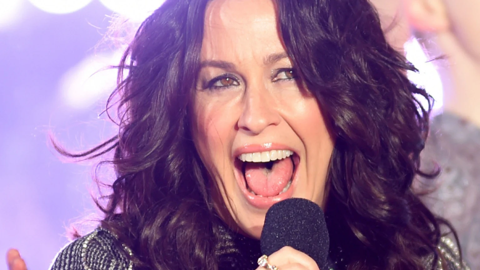 Close-up of Alanis Morissette with dark, wavy hair, singing passionately into a microphone with her mouth wide open, wearing a sparkly top against a blurry purple background.