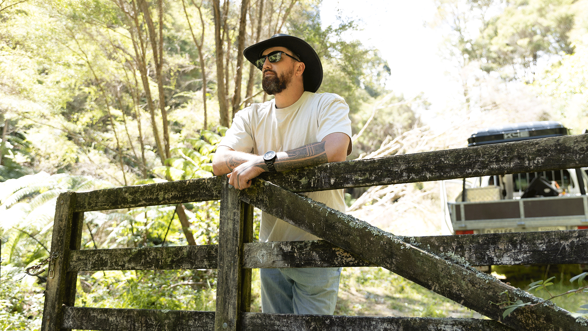 A bearded man in a hat and sunglasses with tattooed arms leans on a weathered wooden fence, with a lush green forest and a distant vehicle in the background.