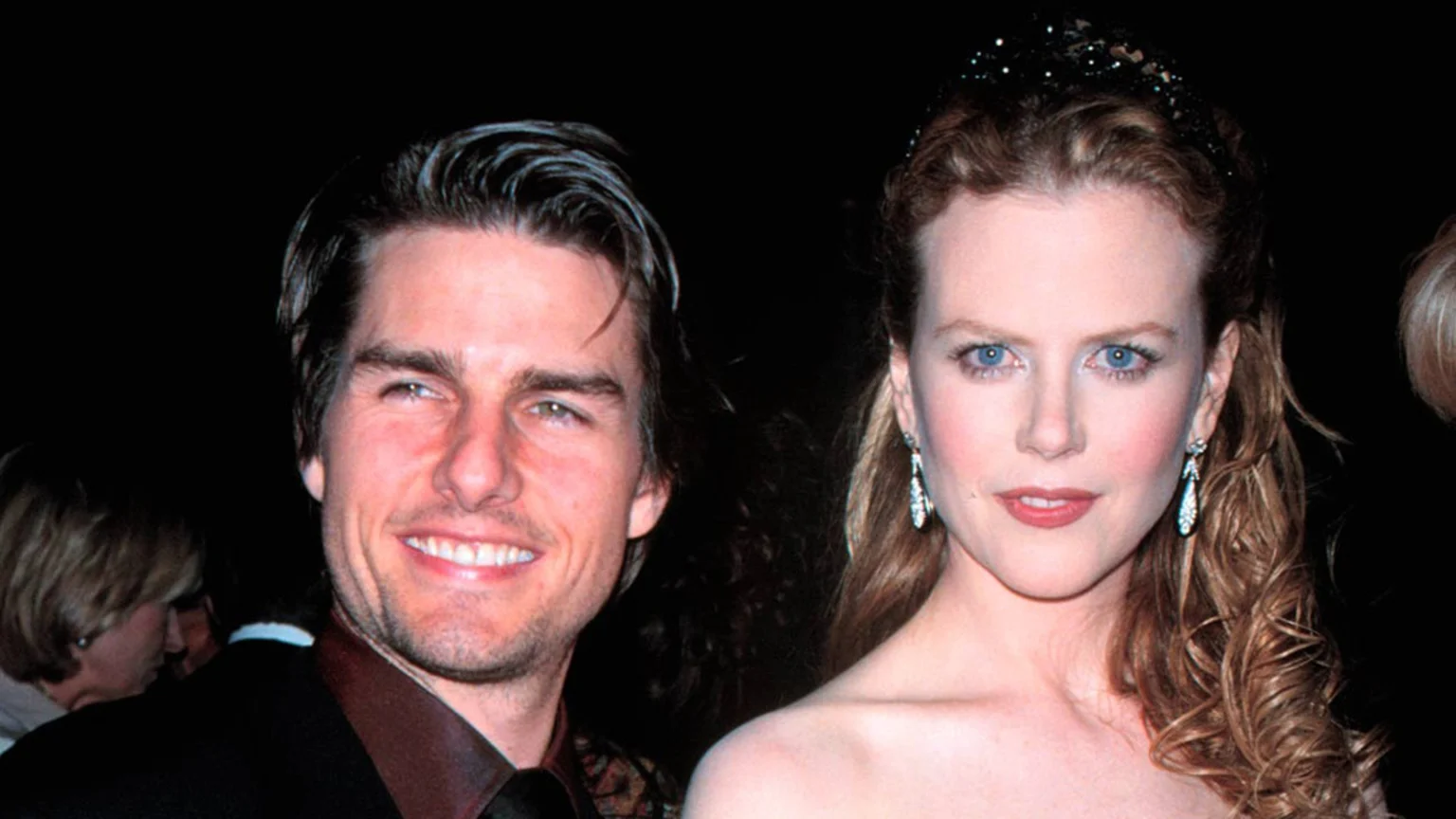 Tom Cruise and Nicole Kidman are pictured together; Cruise smiles in a dark suit, and Kidman looks at the camera with blue eyes, a sparkling headband, and drop earrings.