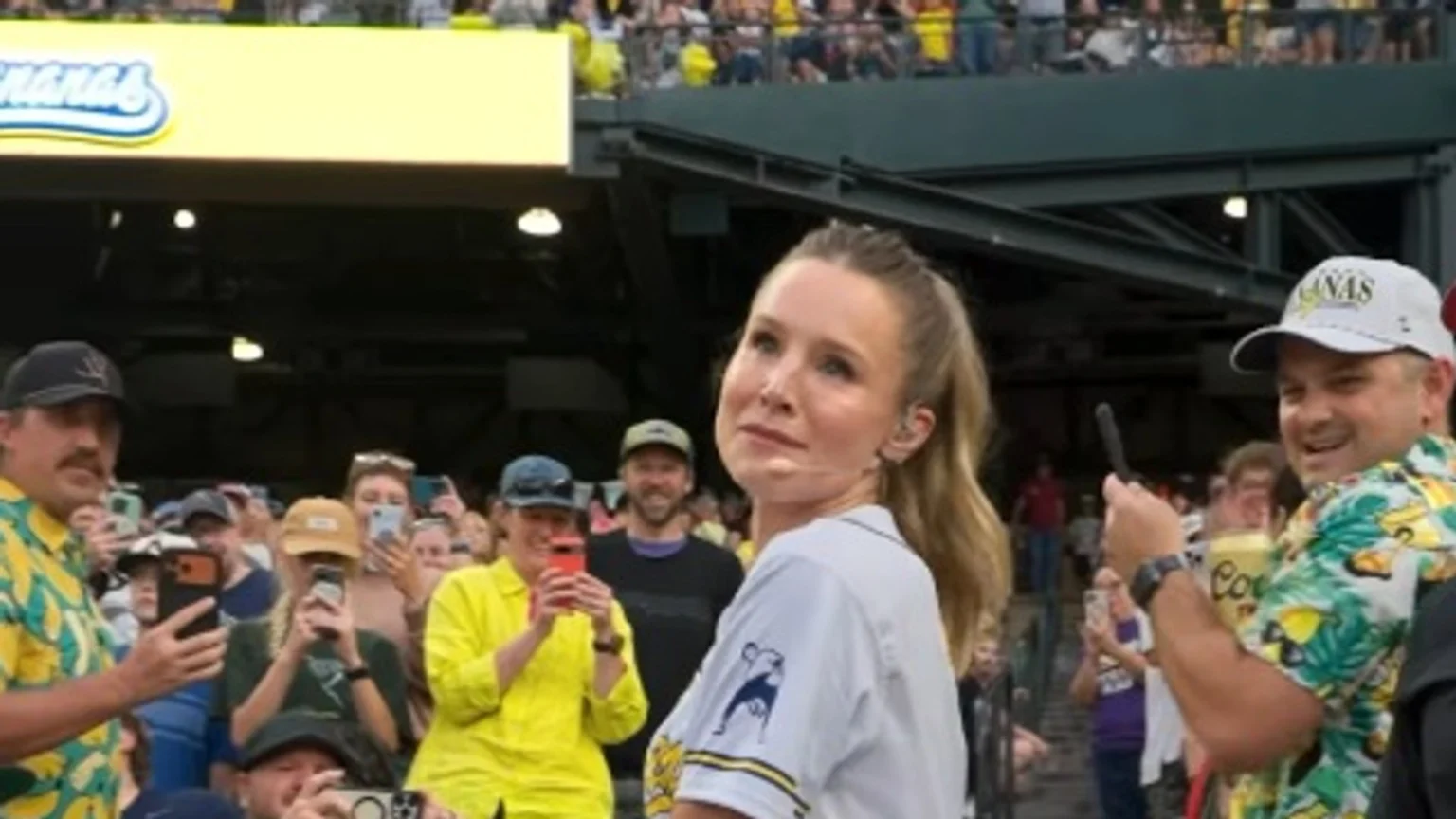 Kristen Bell, wearing a white baseball jersey, looks upward while surrounded by a crowd of fans holding phones, with stadium seating and a yellow "Bananas" sign visible in the background.