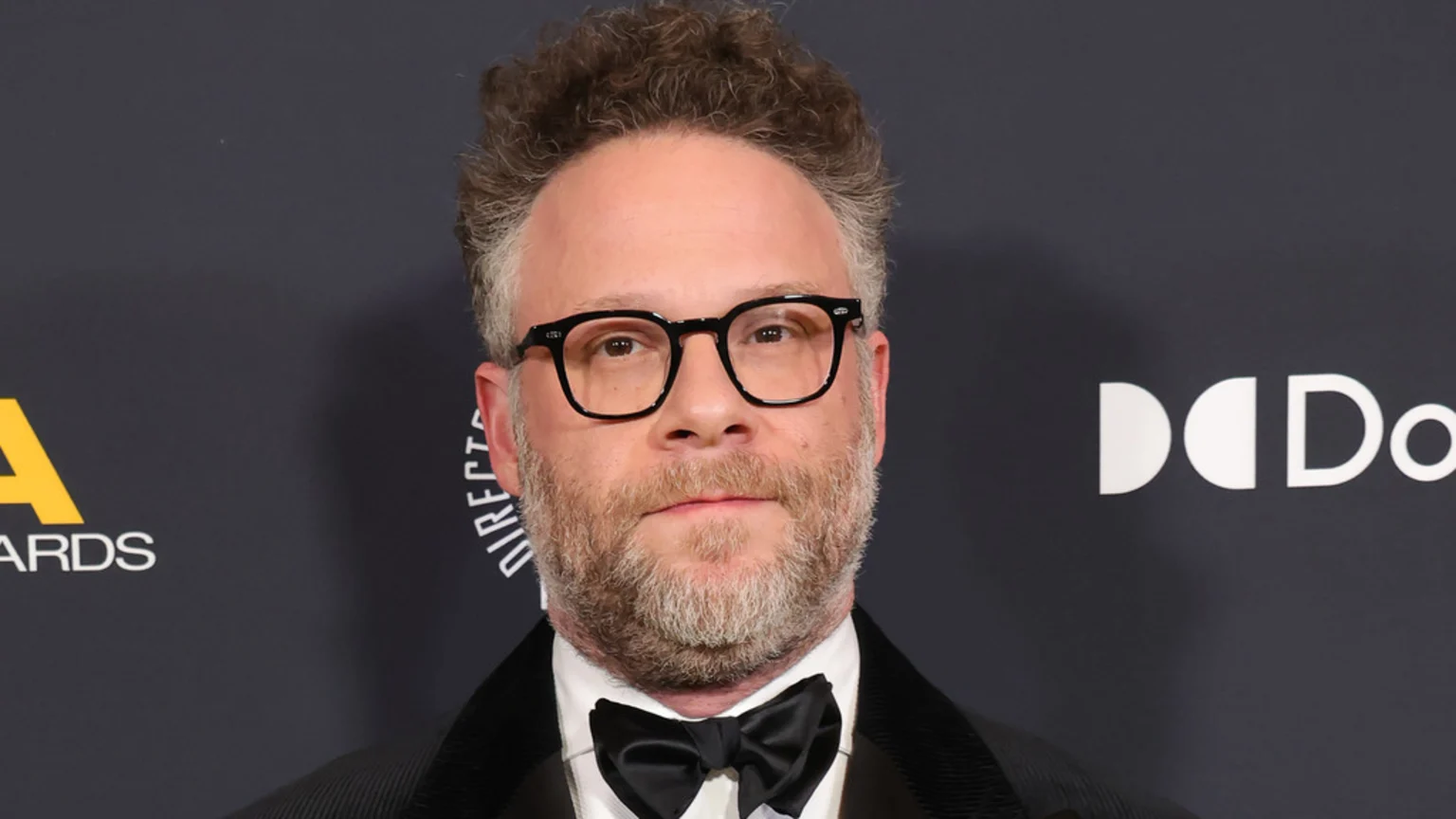Seth Rogen, with curly hair, beard, and black-rimmed glasses, looks directly at the camera while wearing a white shirt and black bow tie, against a dark background with event logos.