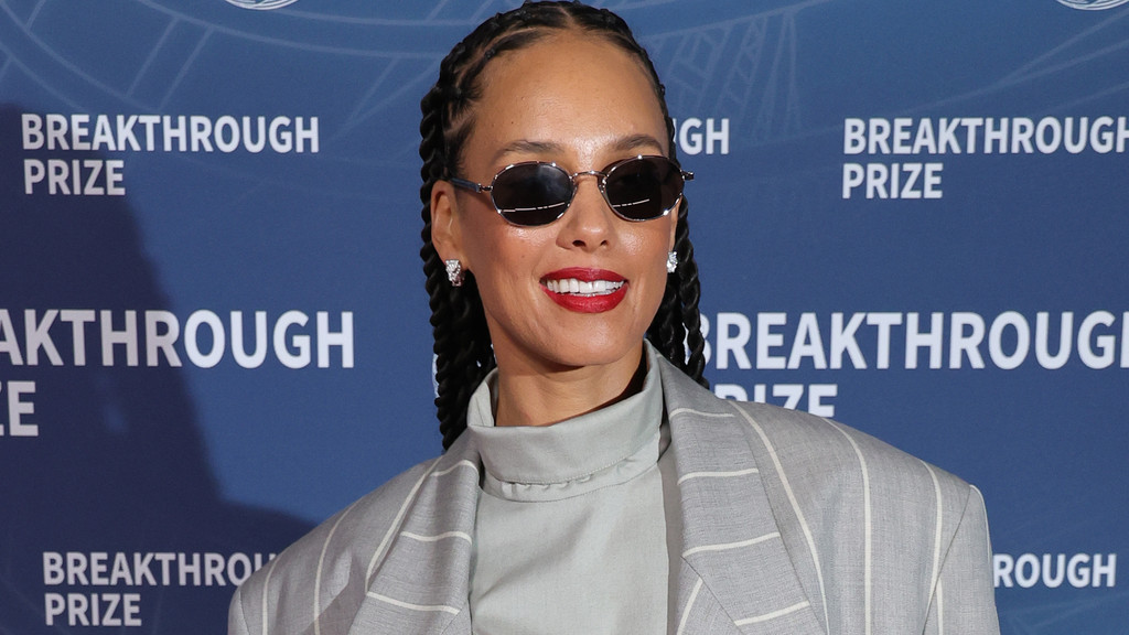 Singer Alicia Keys, with braided hair and sunglasses, smiles wearing red lipstick, a light turtleneck, and a pinstriped blazer in front of a blue "BREAKTHROUGH PRIZE" banner.