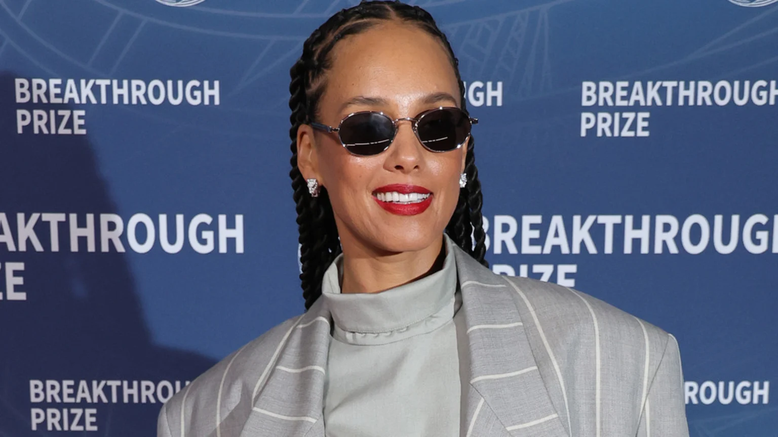 Singer Alicia Keys, with braided hair and sunglasses, smiles wearing red lipstick, a light turtleneck, and a pinstriped blazer in front of a blue "BREAKTHROUGH PRIZE" banner.