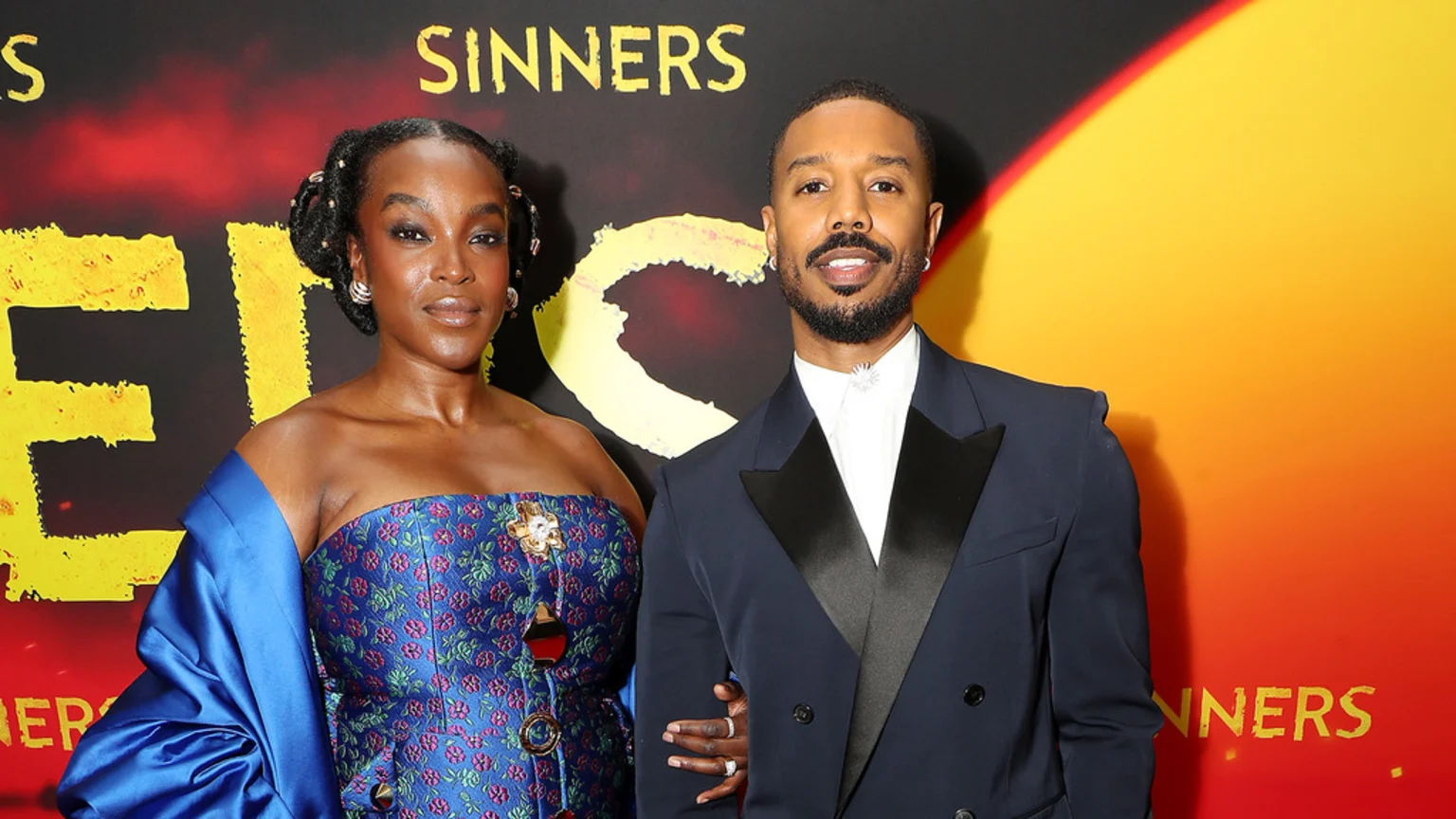 A well-lit indoor portrait shows Michael B. Jordan in a navy blue suit with silver collar pin, standing beside a woman in a strapless blue floral top adorned with gold-toned brooches and accessories. The background features large yellow 'SINNERS' lettering and a vibrant gradient, indicating an event or movie setting.