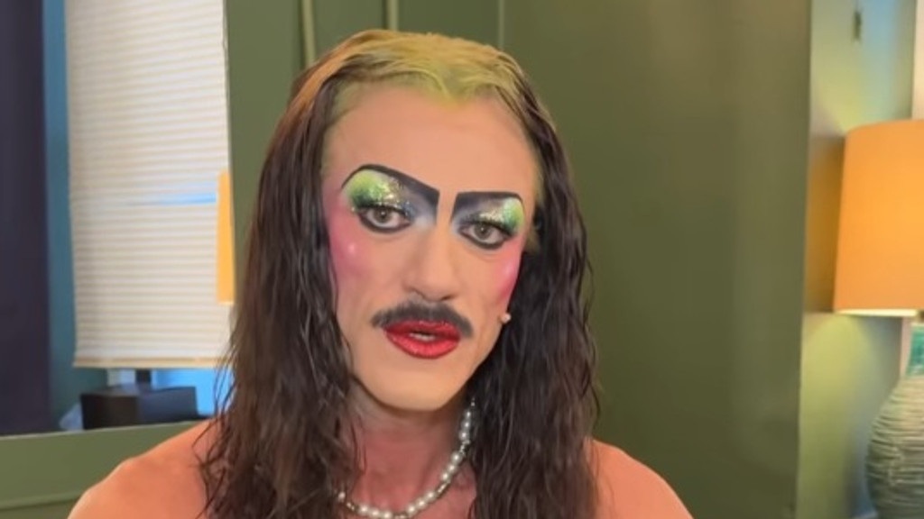 A person in heavy drag makeup, featuring bright green and blue sparkly eyeshadow, bold pink blush, a painted mustache, red lipstick, long dark hair with light green roots, and a pearl necklace, looks directly at the camera.