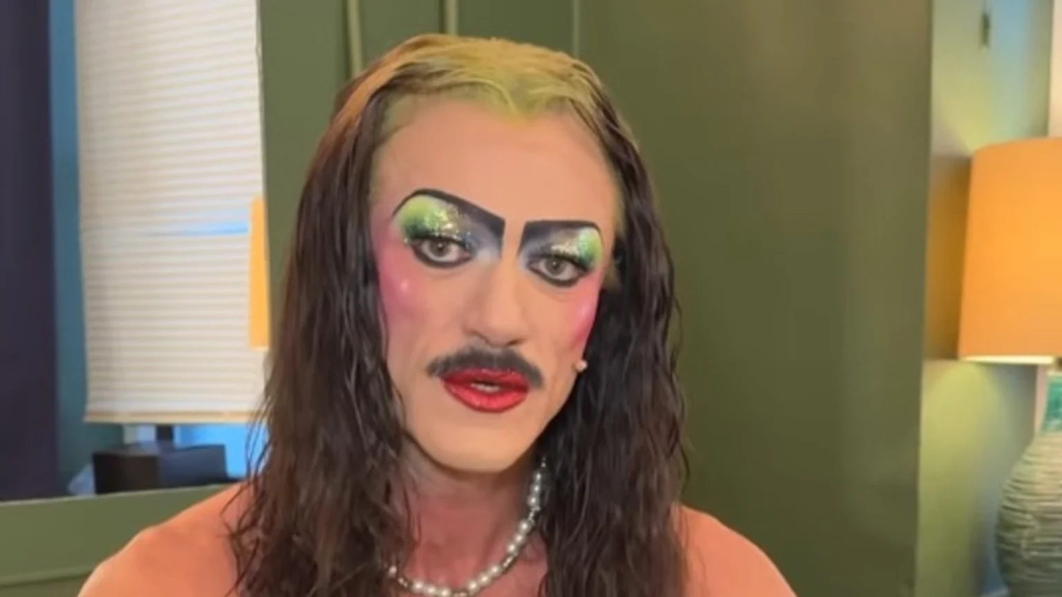 A person in heavy drag makeup, featuring bright green and blue sparkly eyeshadow, bold pink blush, a painted mustache, red lipstick, long dark hair with light green roots, and a pearl necklace, looks directly at the camera.