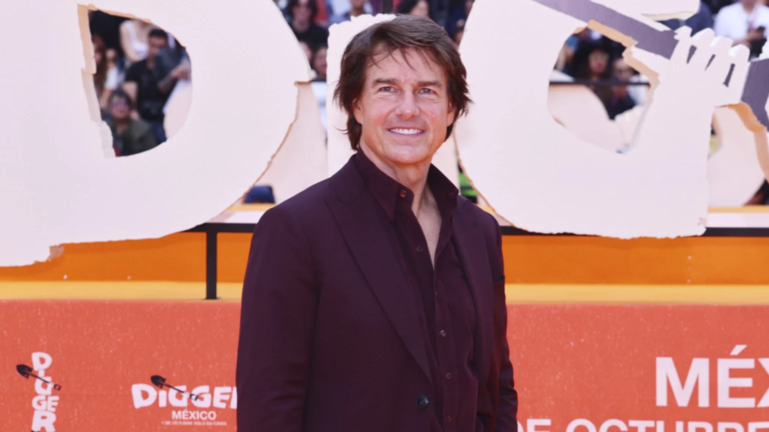Tom Cruise smiles, wearing a dark maroon suit and open-collar shirt, against a background featuring white cutouts and a red wall with "DIGGER MÉXICO" visible.