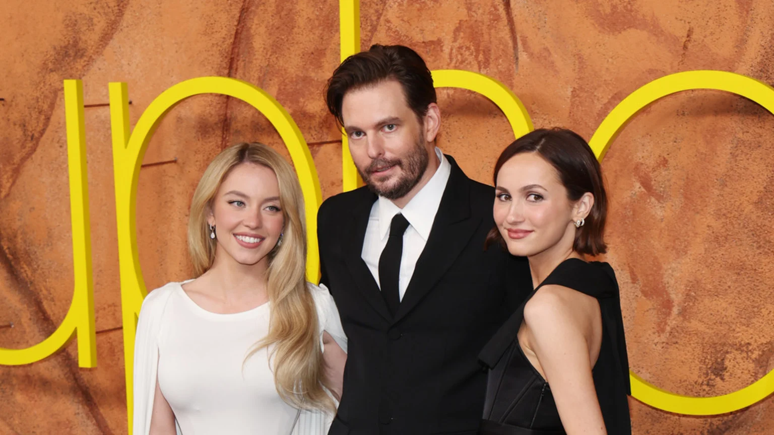 Sydney Sweeney in a white dress smiles between a man in a black suit and a woman in a black dress, posing in front of an orange wall with yellow shapes.