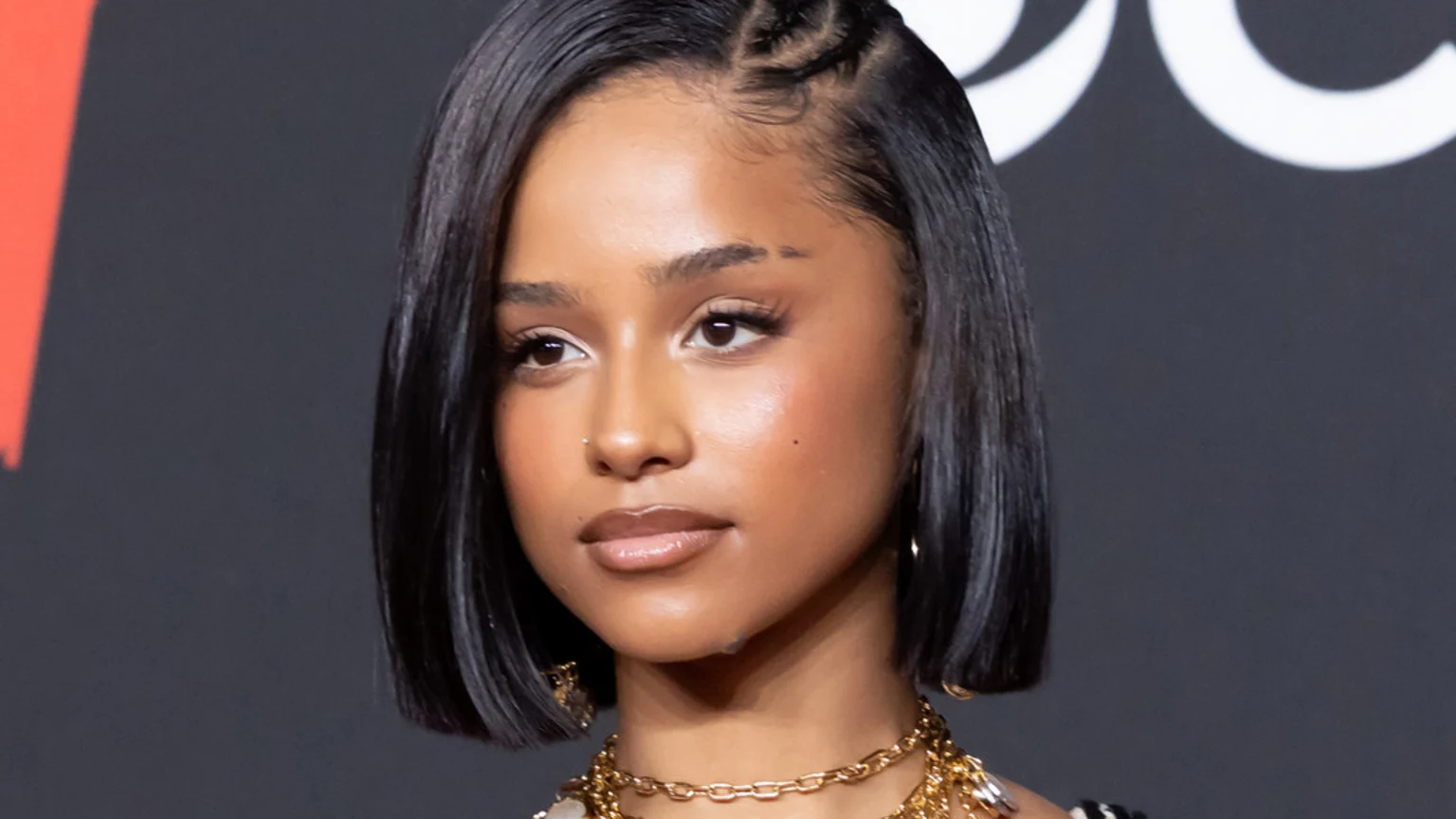 A young Black woman with a bob haircut and braided hairline, wearing a nose piercing and gold necklaces, looks slightly left against a dark background with abstract shapes.