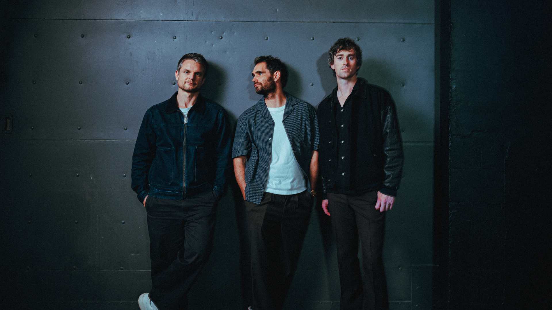 Three men stand against a dark wall, two looking at the camera, one looking to the side. They are casually dressed in jackets, shirts, and trousers.