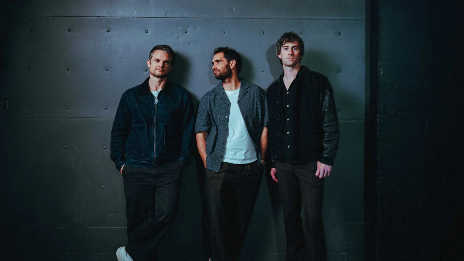 Three men stand against a dark wall, two looking at the camera, one looking to the side. They are casually dressed in jackets, shirts, and trousers.