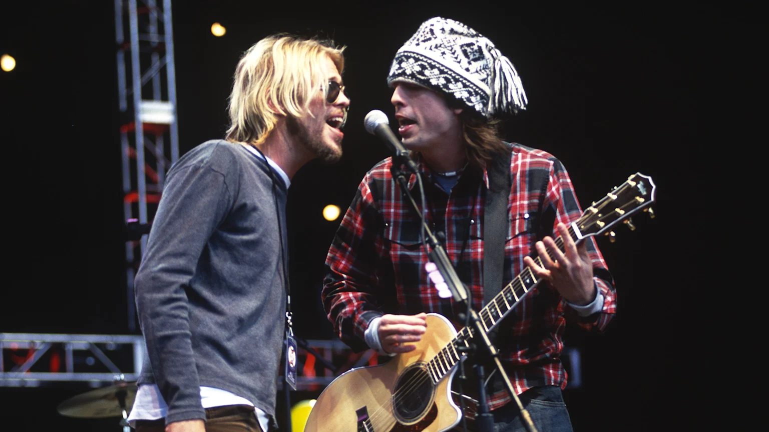 Two musicians sing into a shared microphone on stage: a blond man with a beard and sunglasses, and another in a patterned knit hat and plaid shirt playing an acoustic guitar.