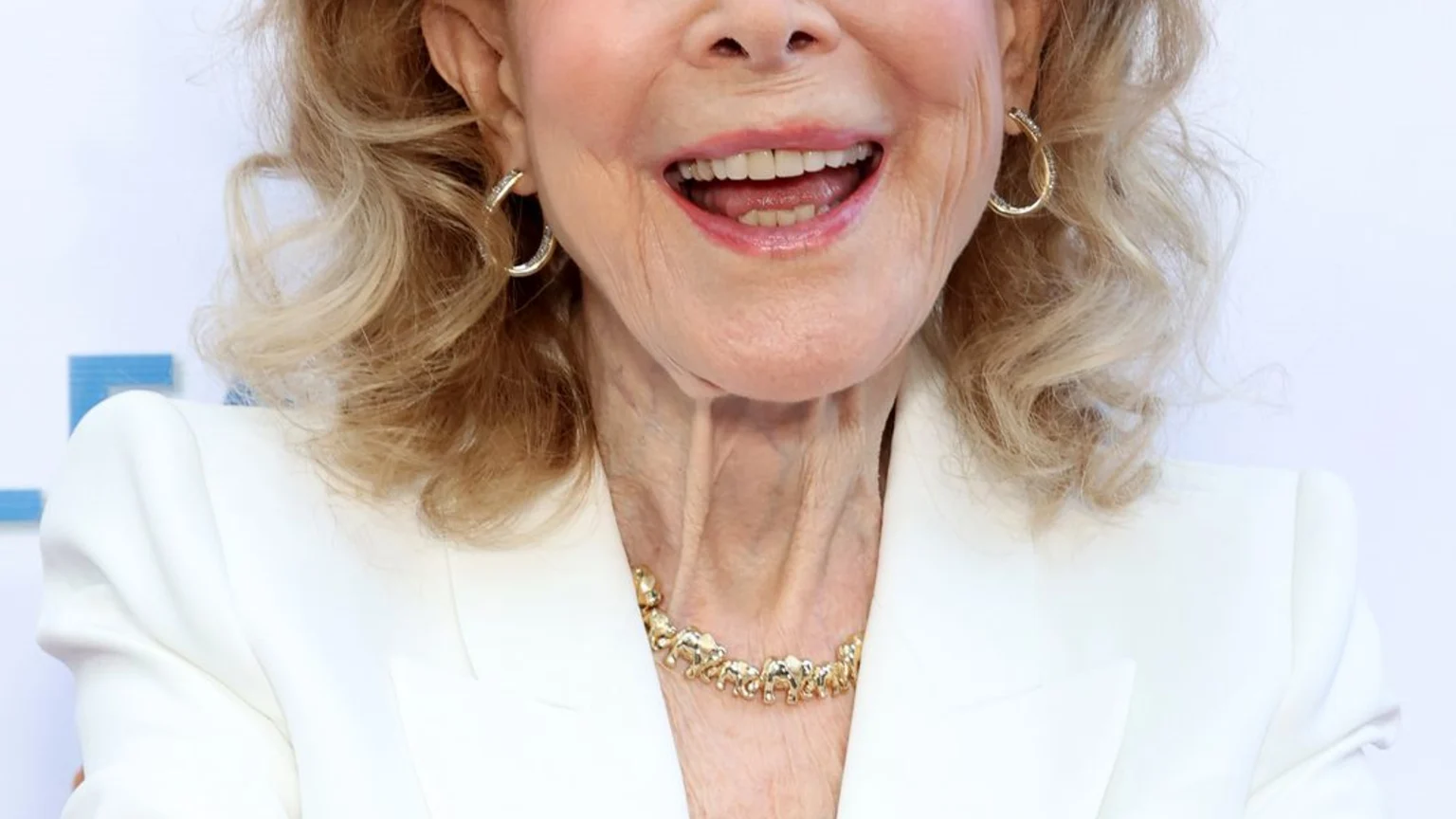 Barbara Eden, an older woman with blonde hair, smiles broadly, wearing blue eyeliner, gold hoop earrings, a gold elephant necklace, and a white blazer.