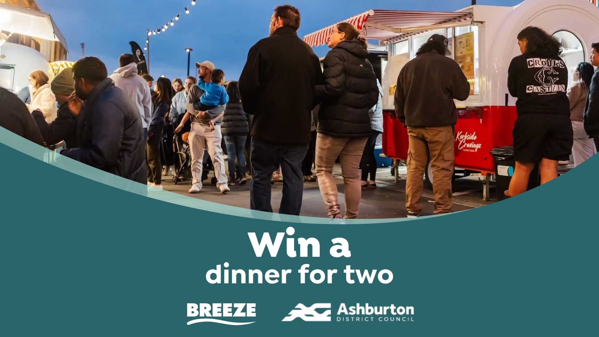 A bustling night scene shows many people gathered around food trucks, with a teal overlay at the bottom promoting "Win a dinner for two" from Breeze and Ashburton District Council.