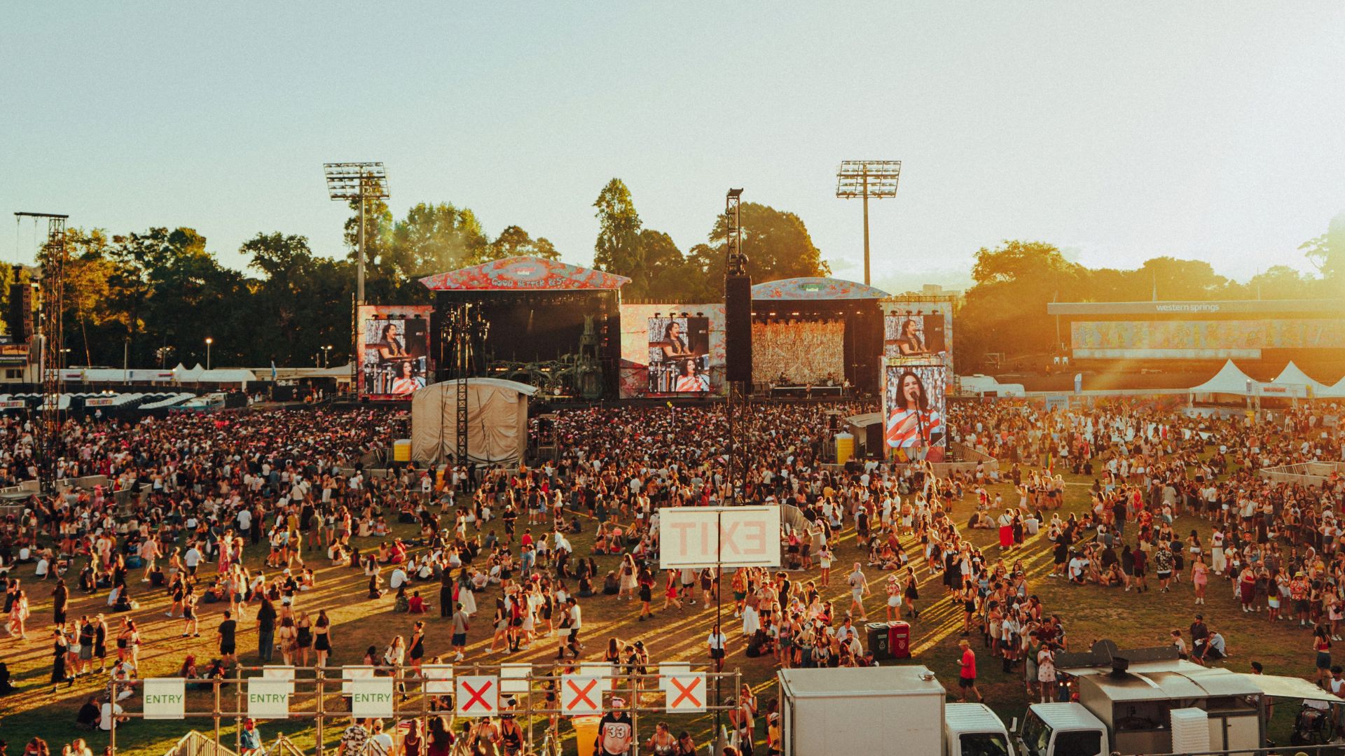 A crowded outdoor music festival at sunset, with a large audience gathered before multiple stages displaying performers on screens, and 'Western Springs' visible on a background building.