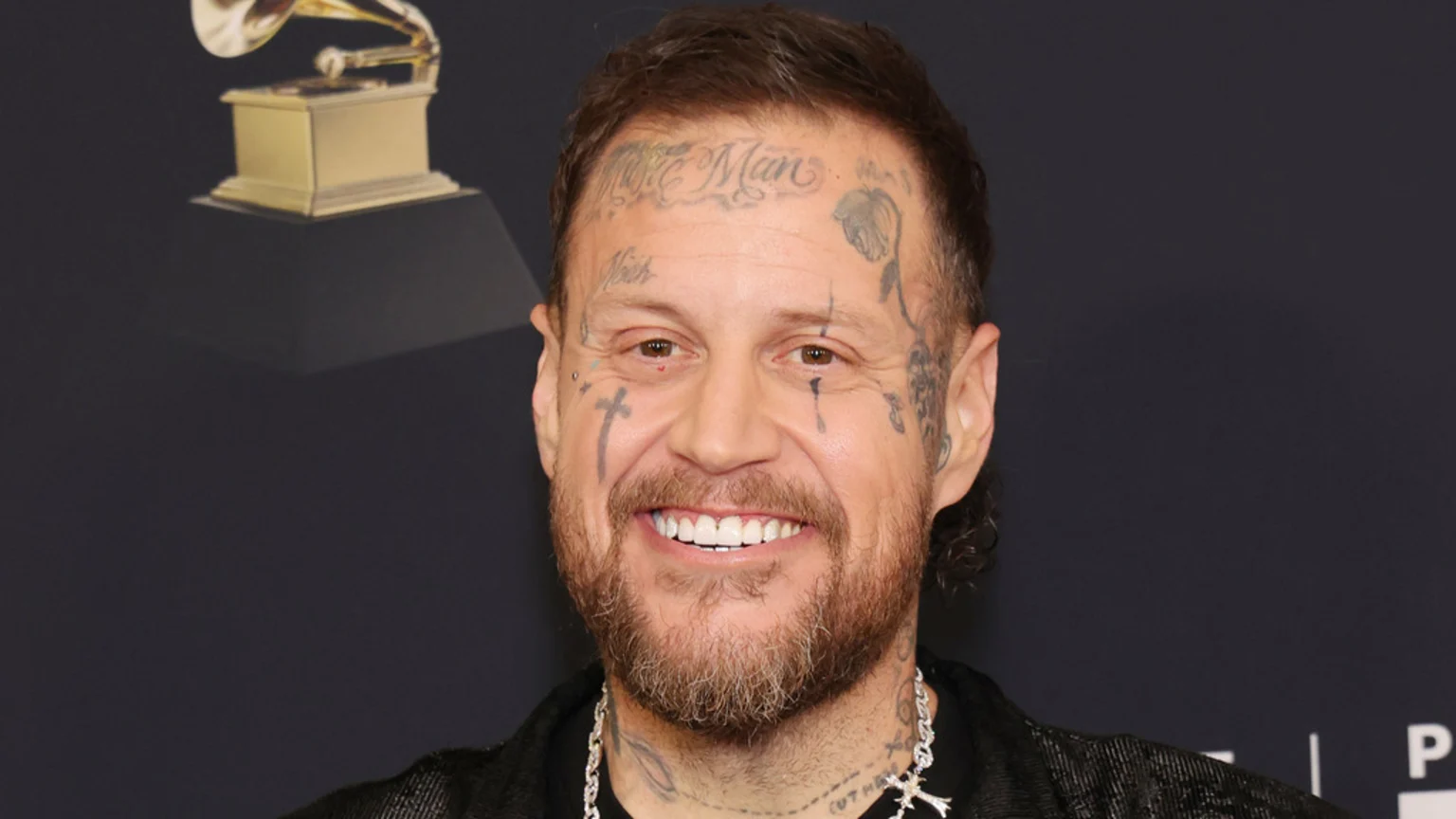 A smiling man with a beard and numerous facial tattoos, including "More Man" on his forehead and crosses on his face, stands in front of a Grammy award.