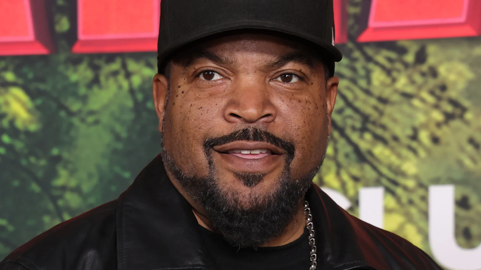 Close-up of Ice Cube wearing a black cap and leather jacket, smiling gently with a blurred green and red background.