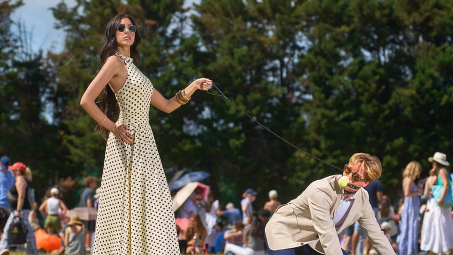 A woman in a polka-dot dress and sunglasses holds a leash connected to a man on all fours, wearing a suit jacket, sunglasses, and a tennis ball gag, outdoors in a park.