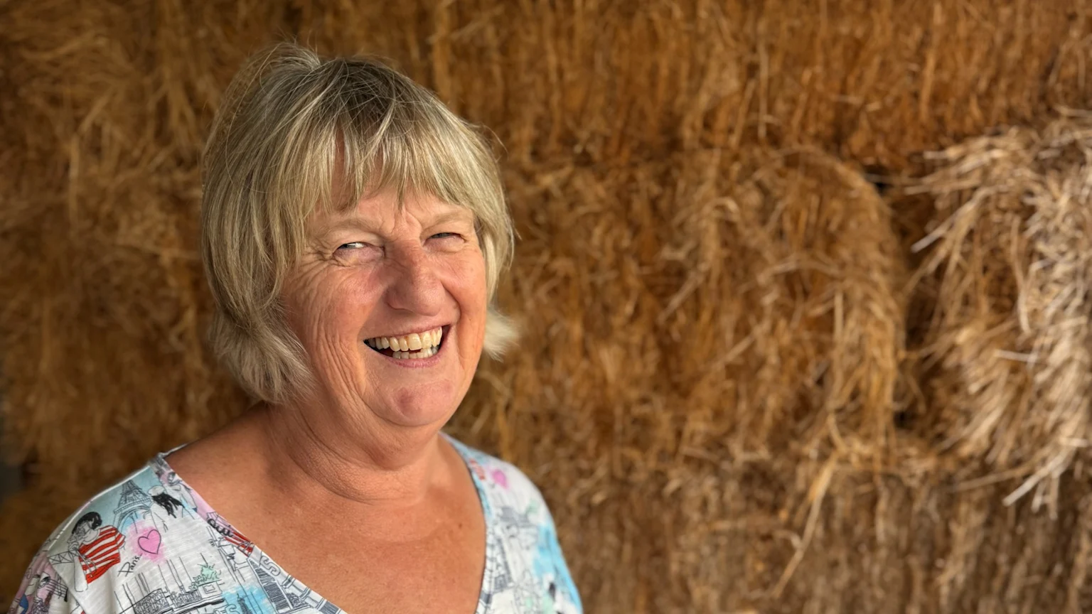 A blonde woman smiles broadly, wearing a patterned top, in front of a blurred background of hay bales.