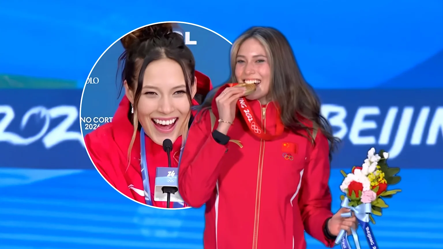 Eileen Gu, wearing a red athletic jacket with the Chinese flag and Olympic rings, joyfully bites her gold medal and holds a floral bouquet at an indoor awards ceremony. She is also shown speaking into a microphone, with event branding for Beijing 2022 visible in the background.