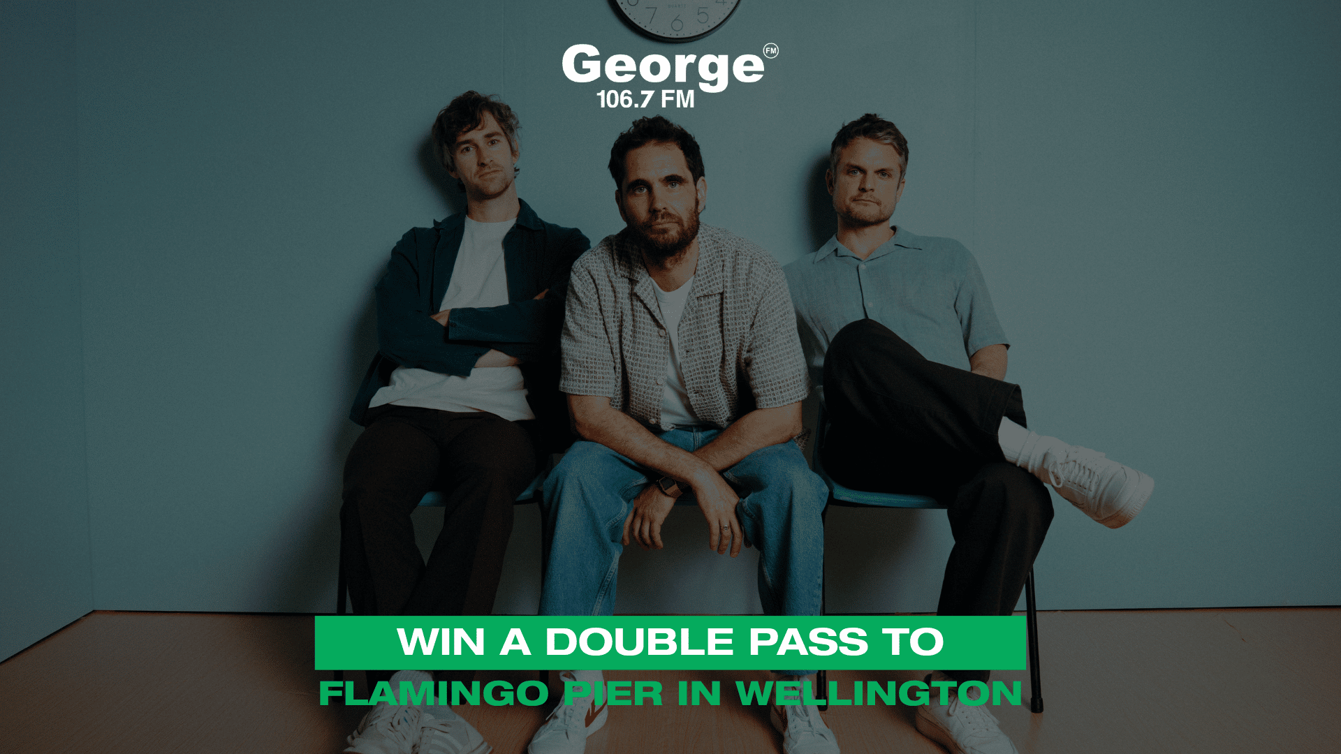 Three men sit against a teal wall, with a "George 106.7 FM" logo and clock visible above, and a green banner reading "WIN A DOUBLE PASS TO FLAMINGO PIER IN WELLINGTON" below.