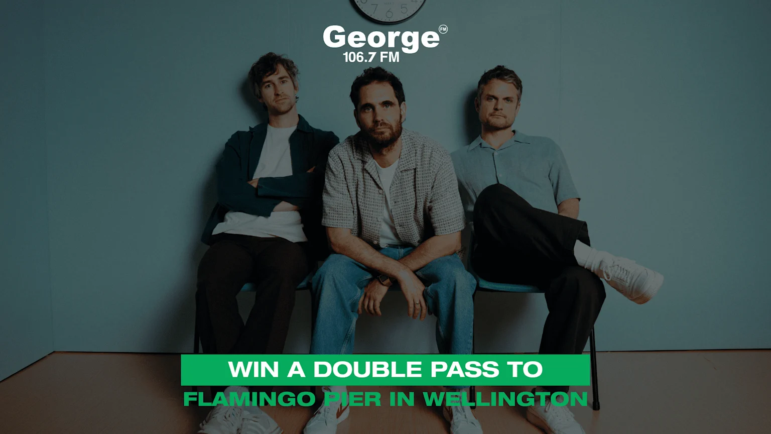 Three men sit against a teal wall, with a "George 106.7 FM" logo and clock visible above, and a green banner reading "WIN A DOUBLE PASS TO FLAMINGO PIER IN WELLINGTON" below.