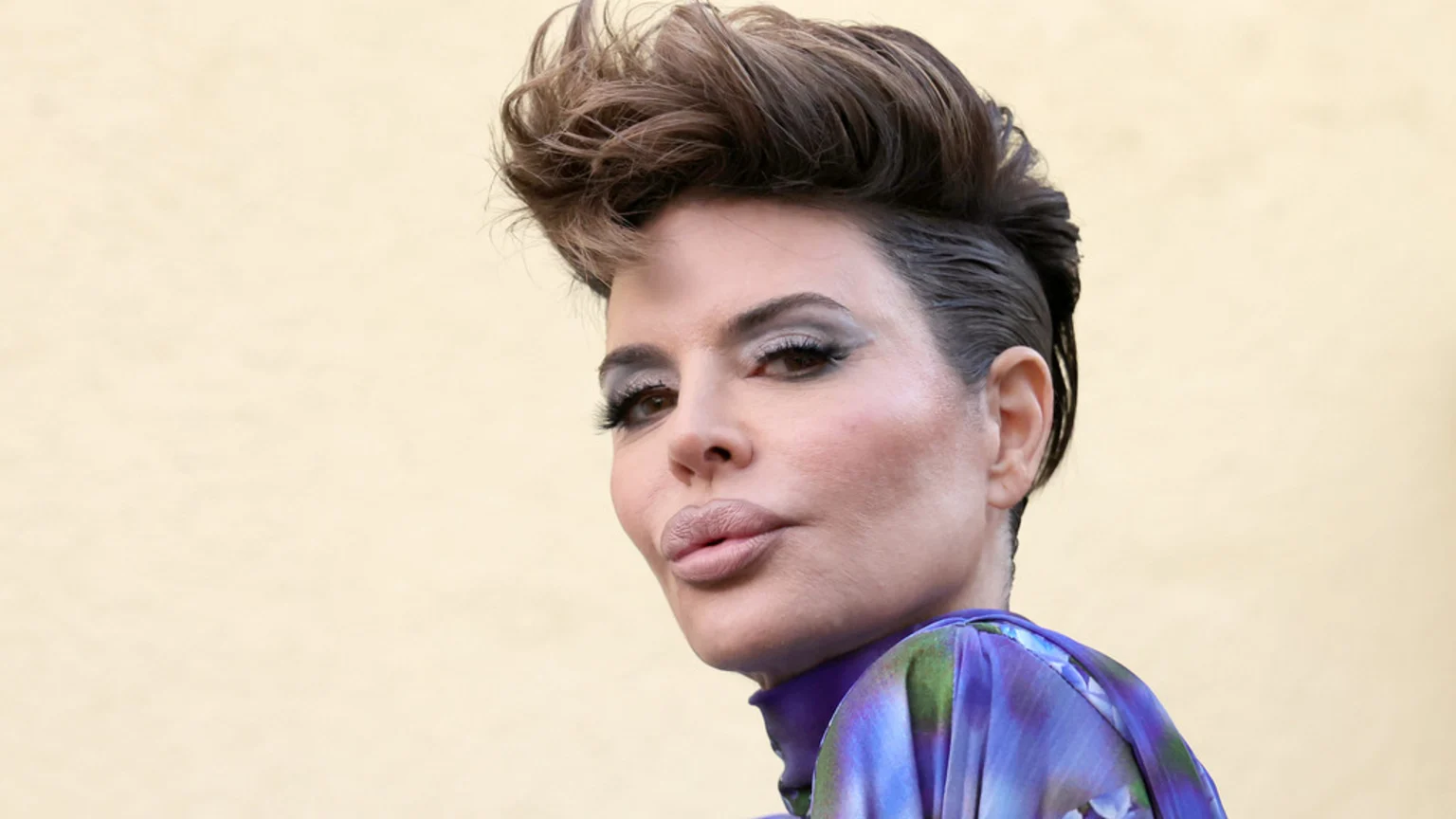 Close-up of a woman with dark hair styled in a high pompadour and shaved sides, full lips, and heavy eye makeup, wearing a purple and green patterned top.