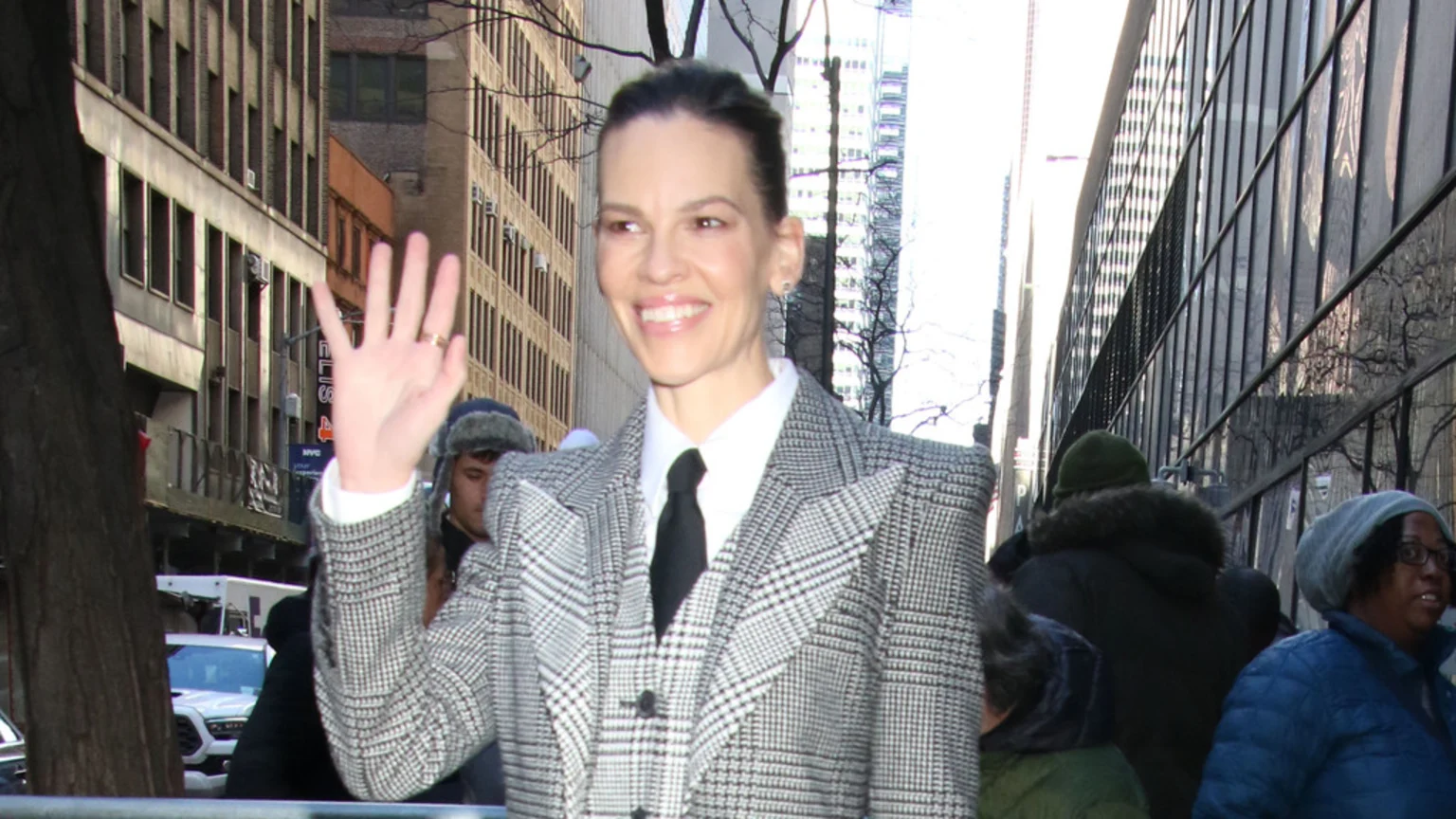 Hilary Swank smiles and waves, wearing a grey plaid suit with a white shirt and black tie, on a bustling city street with buildings and blurred people in the background.