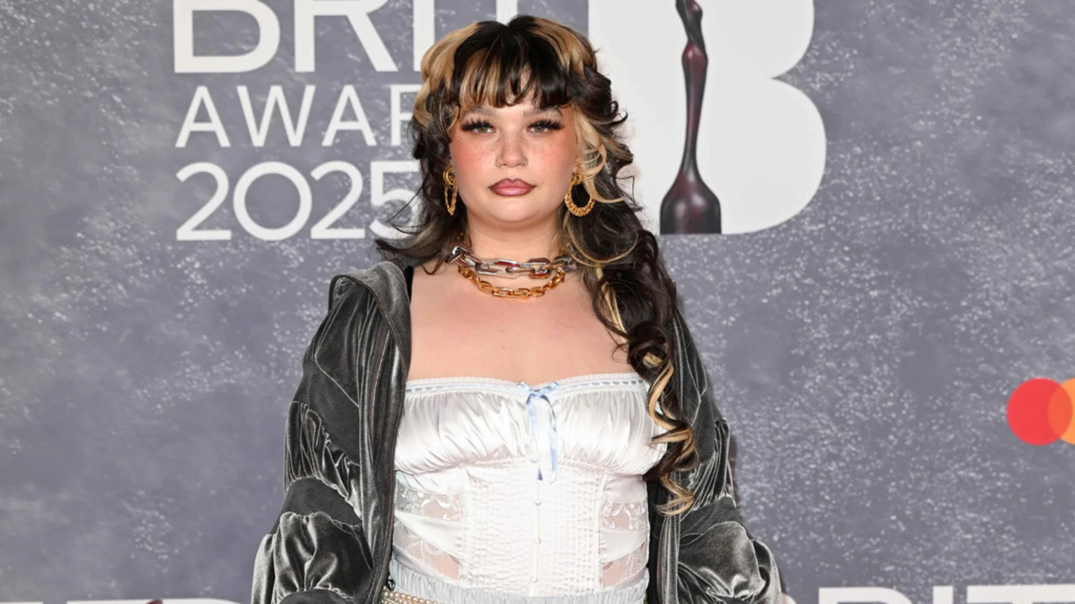 A young woman with dark hair and blonde streaks, heavy makeup, and a nose piercing, wearing a chunky necklace, white corset top, and velvet jacket, poses in front of a 'BRIT AWARDS 2025' sign.